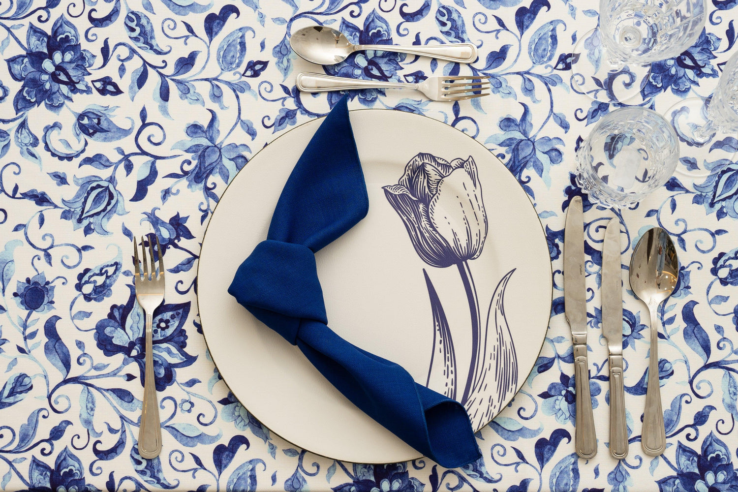 A table flatlay in blue with a printed tablecloth and blue napkin with silverware.