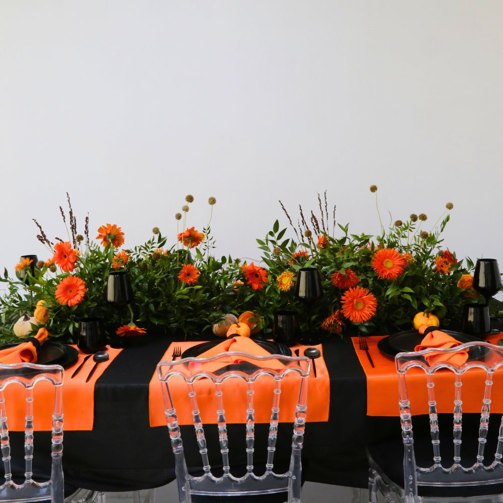 Pumpkins & Petals- Dining Setup with a black tablecloth, orange accents, clear chairs, and a vibrant floral centerpiece, perfect for an elegant dinner party for eight.