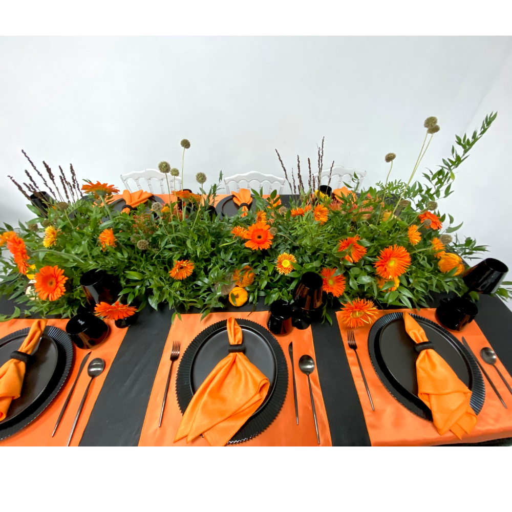 Pumpkins & Petals- Dining Setup: Elegant tablescape featuring lush foliage, vibrant orange flowers, black dinnerware, and modern flatware, set for eight guests with sleek white chairs.