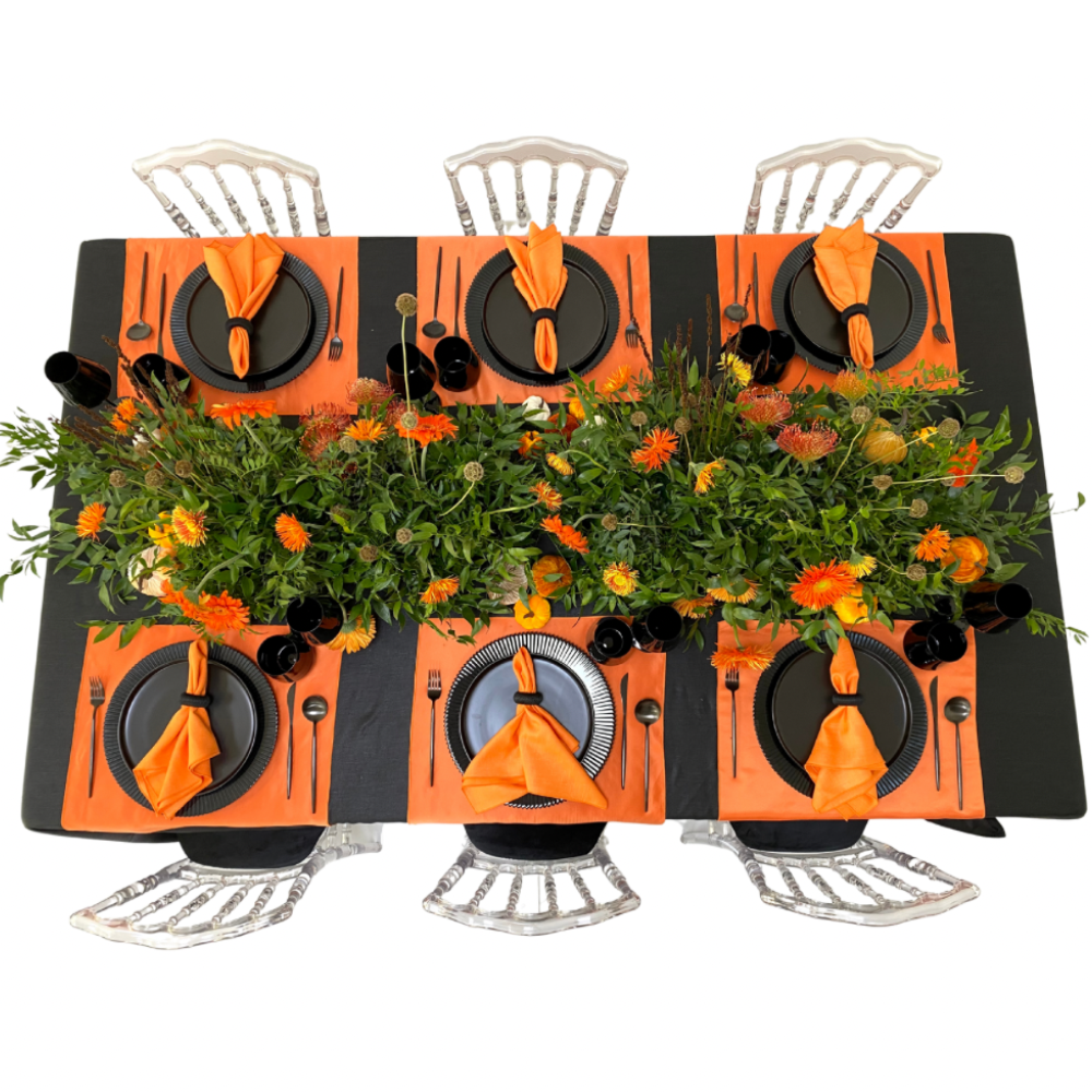 Pumpkins & Petals- Dining Setup: Overhead view of a table for six with orange accents, greenery centerpiece, black dinnerware, and clear chairs, ideal for autumn events.