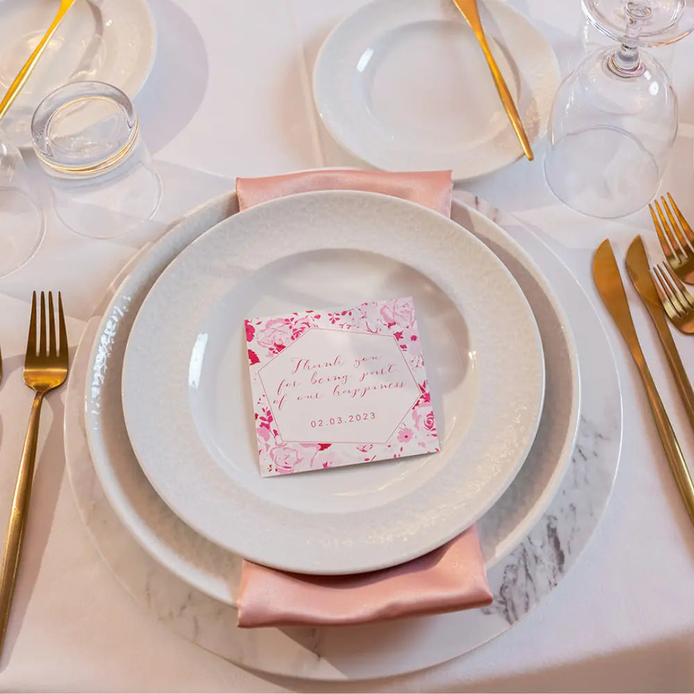 Elegant setting featuring the CHARGER PLATE - MARBLE TRAY, adorned with a blush satin napkin and floral card, complemented by gold-tone flatware and glasses.