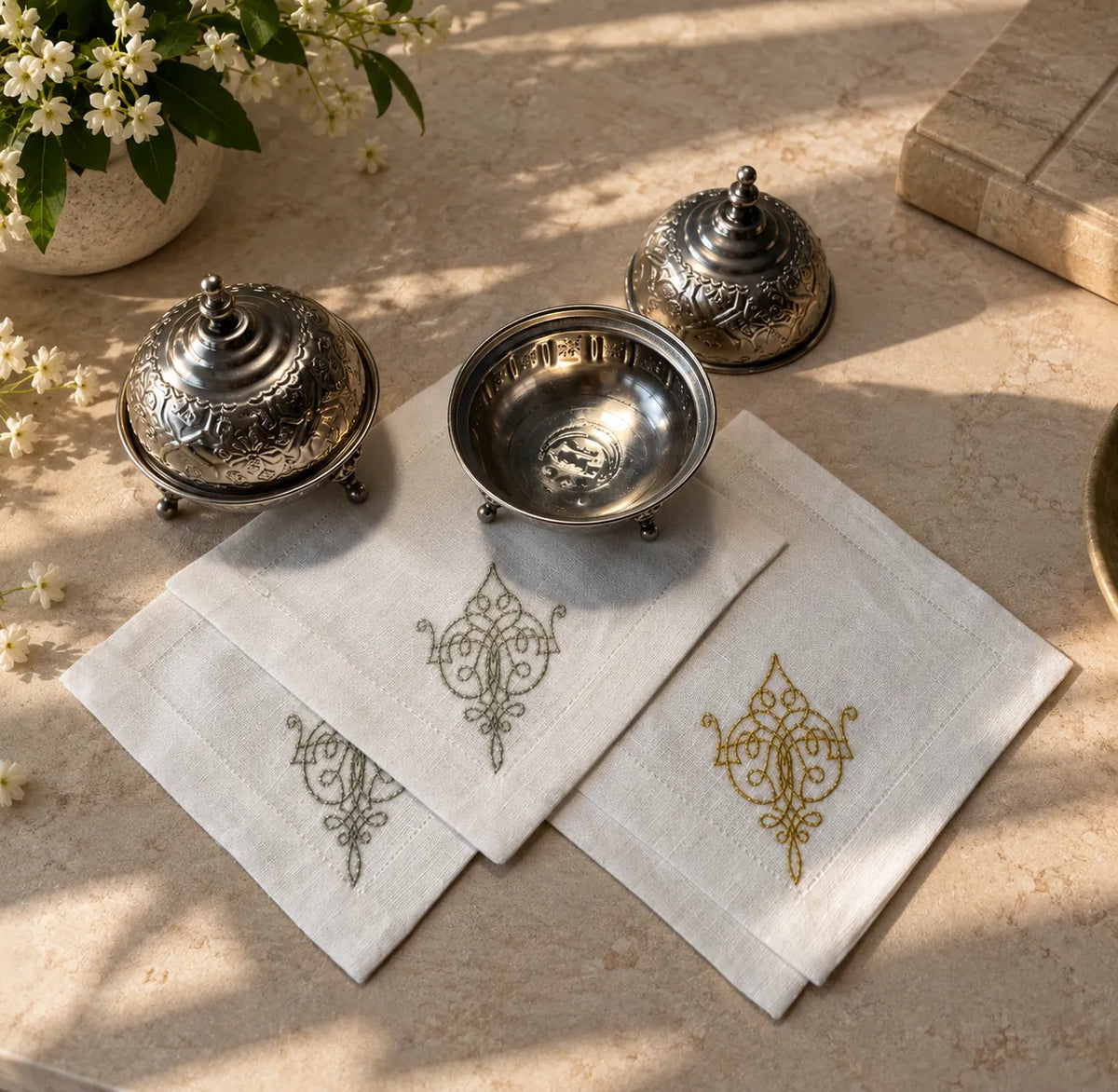 Three white embroidered cotton napkins with ornate gold and gray motifs, arranged on a beige stone surface alongside decorative silver metal dishes and small white flowers.