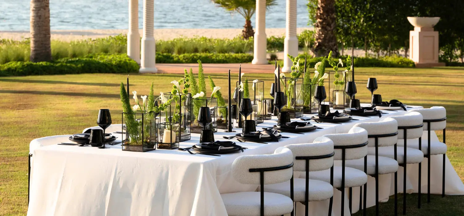 Elegant outdoor dining setup with white tablecloths and black tableware on a grassy area by the beach.