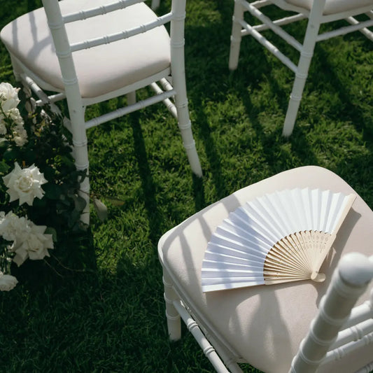 Cluster of White Chiavari Chairs on lawn with open hand-fan and flowers, showcasing elegant event seating from Party Social.