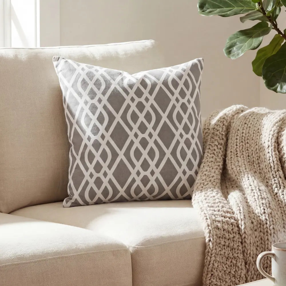 CUSHION GREY CURSIVE PRINT on a beige sofa, paired with a chunky-knit throw and a houseplant, creating a cozy, neutral living-room setting.