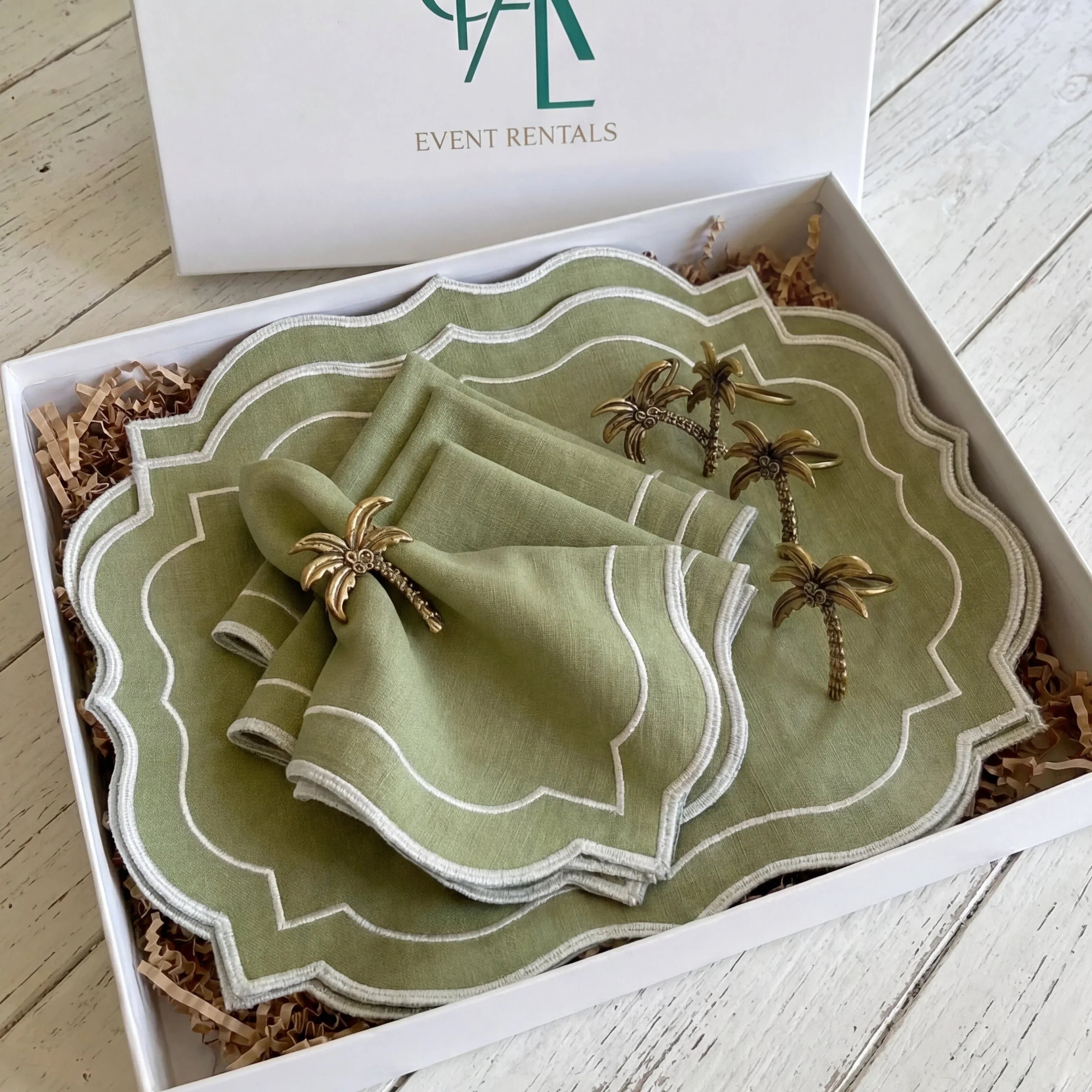 Palm Serenity Gift Set: A gift box featuring green linen placemats, matching napkins, and brass palm-tree napkin rings, nestled in shredded packing paper.