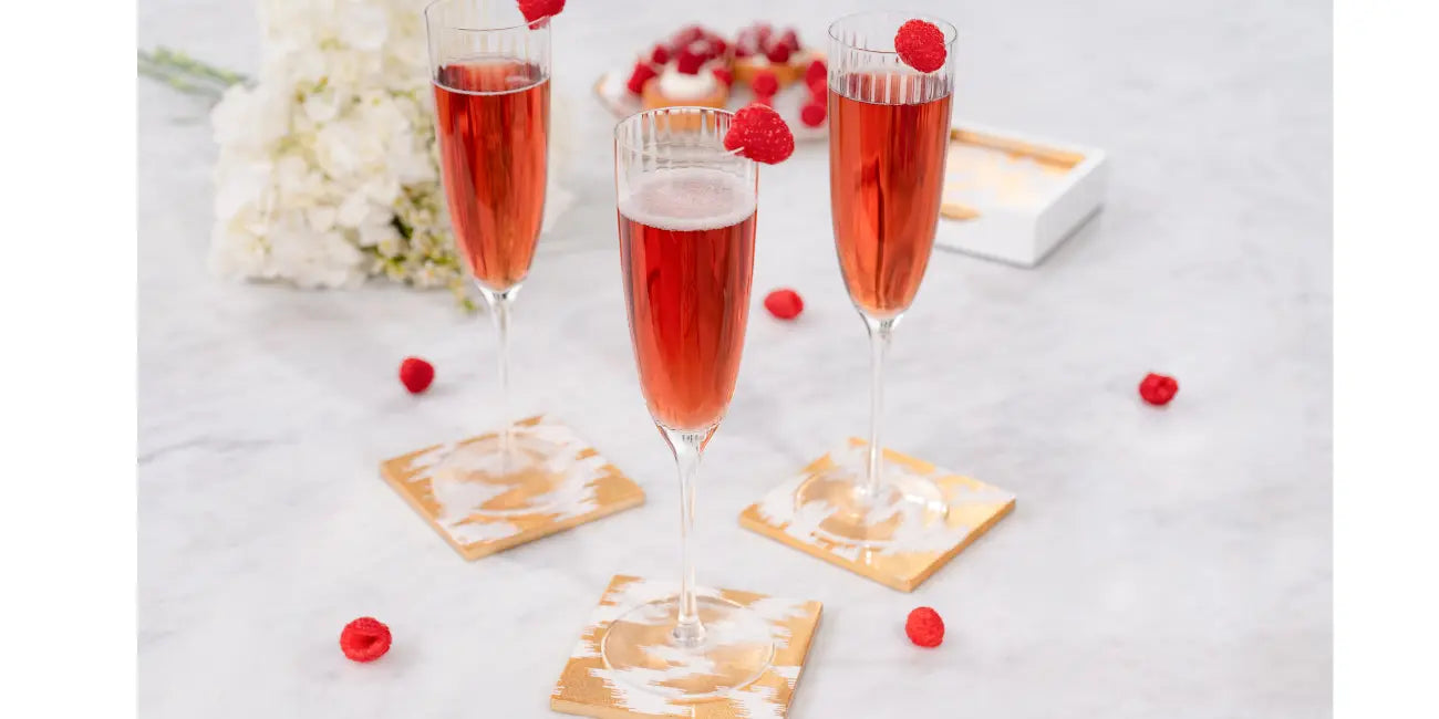 Three glasses of pink sparkling wine glass in a golden print coaster with raspberries on a light gray surface with white flowers.