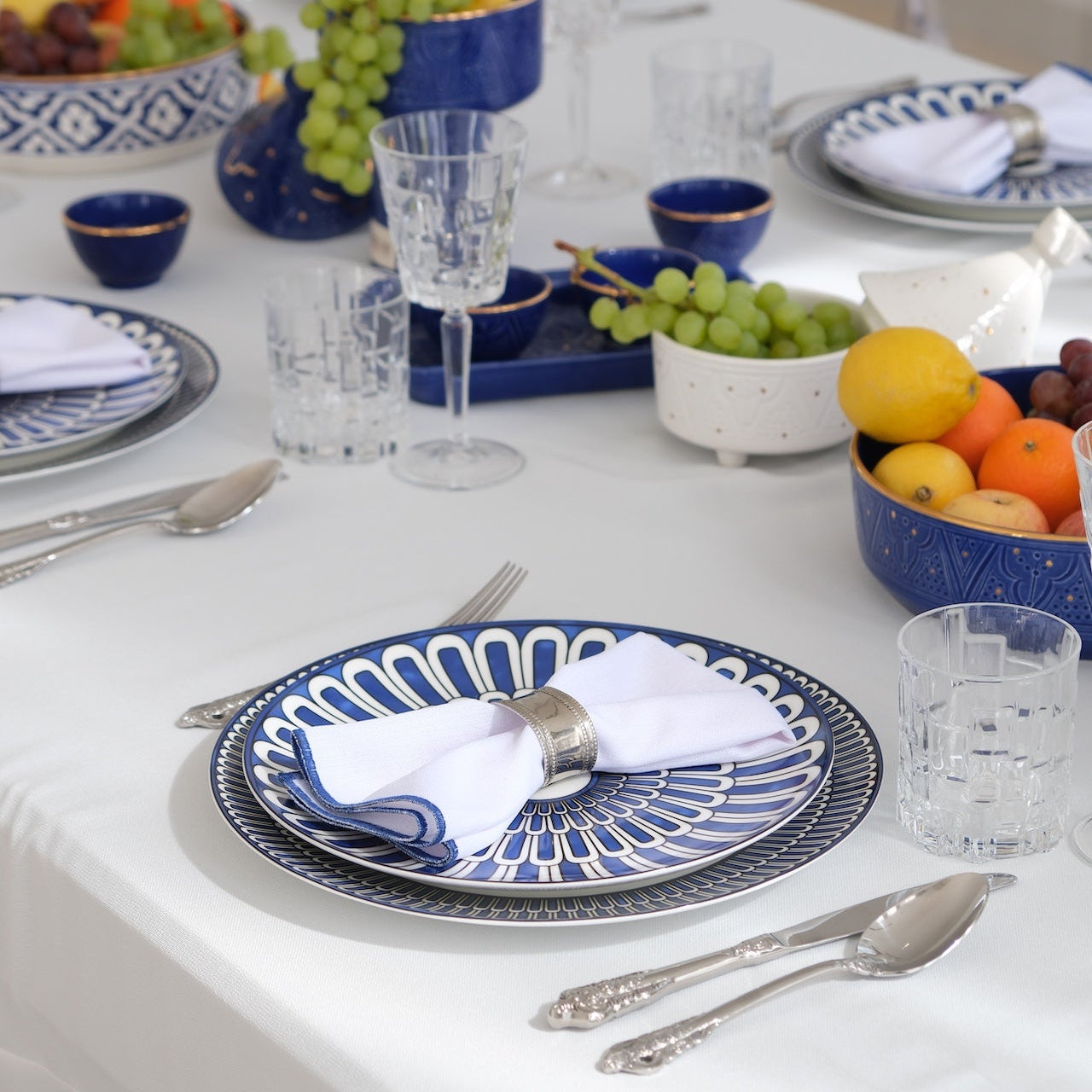 Qamar Bleu - Dinner Setup: A formal table for eight, featuring blue-and-white plates, silver napkin rings, crystal glassware, gold cutlery, fruit centerpieces, and elegant candle holders.