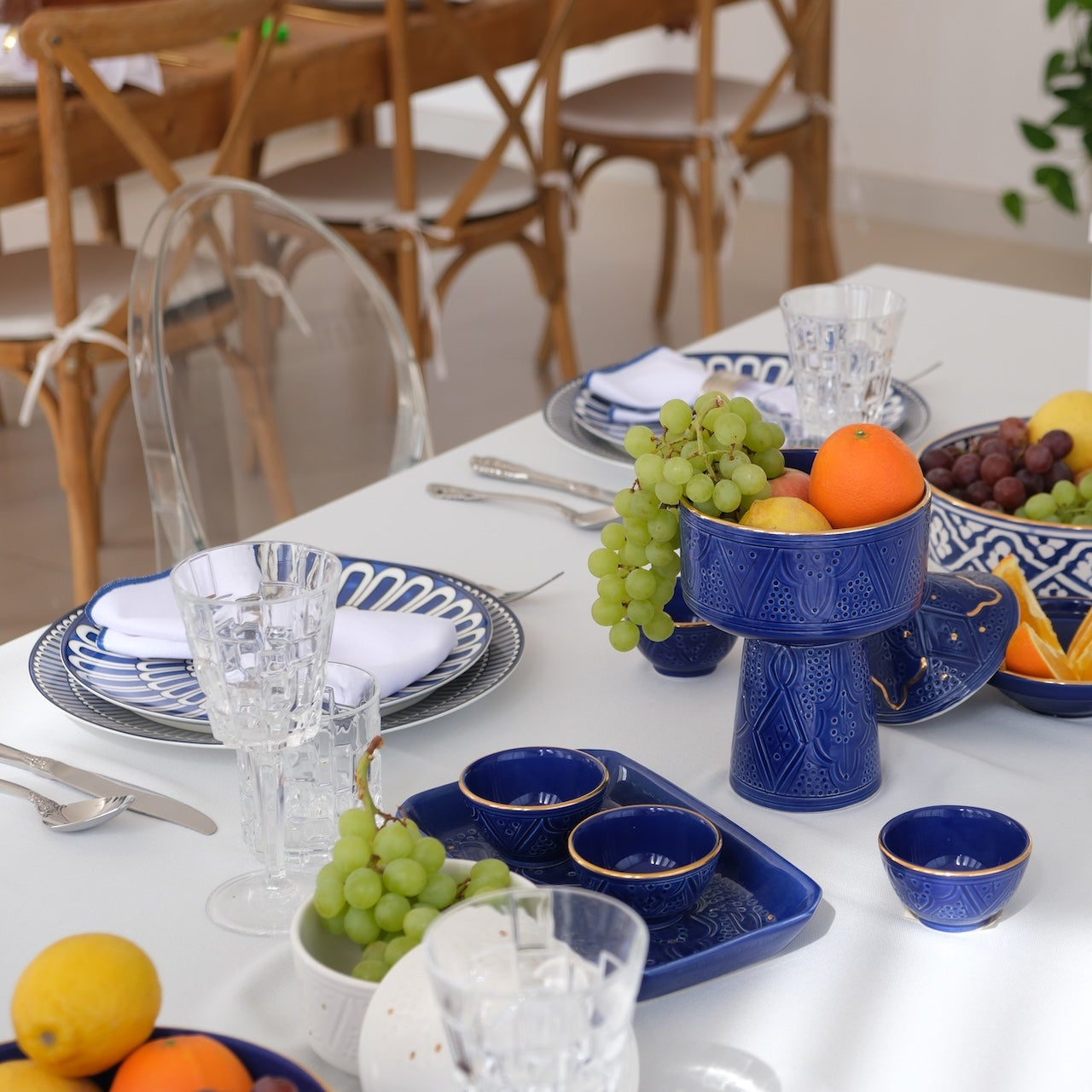 Qamar Bleu - Dinner Setup: Elegant dining table set for eight with blue-and-white ceramic plates, crystal glasses, silverware, and cobalt-blue serving pieces filled with fruit.