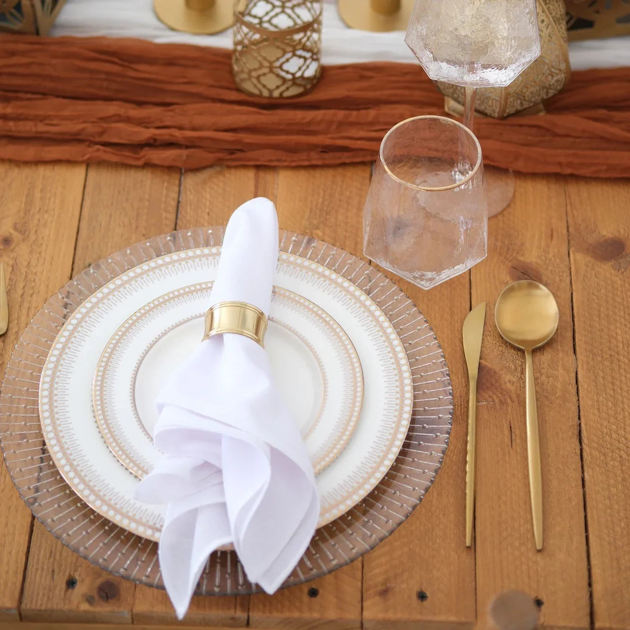 The Noor Royale - Dinner Setup featuring elegant plates, gold flatware, faceted glasses, napkins, and candles on a wooden table, ideal for refined gatherings.