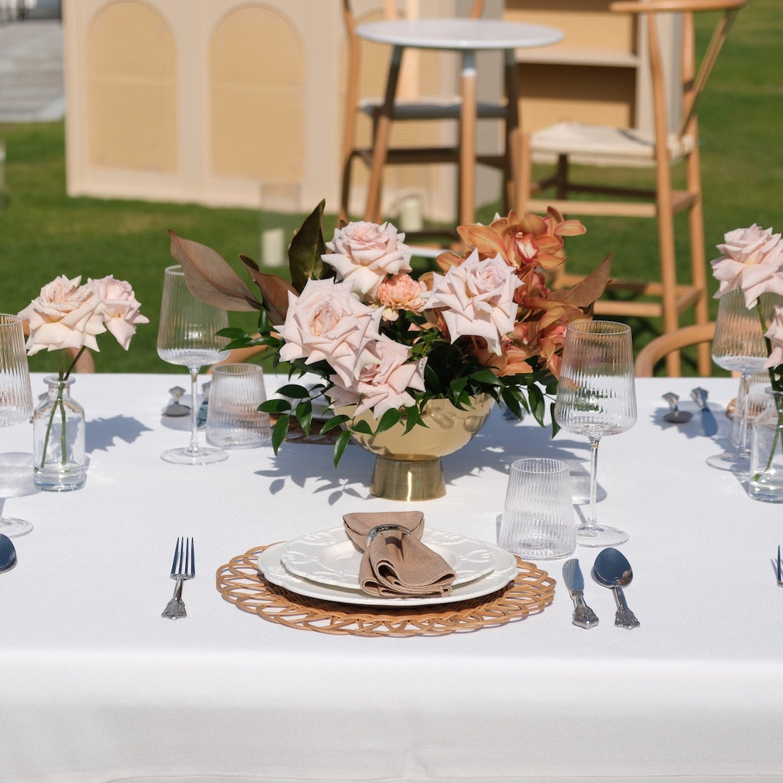 Luxe Alfresco - Dinner Setup featuring a wooden table, rattan chargers, elegant glassware, blush roses, and crossback chairs for eight guests. Perfect for upscale outdoor gatherings.