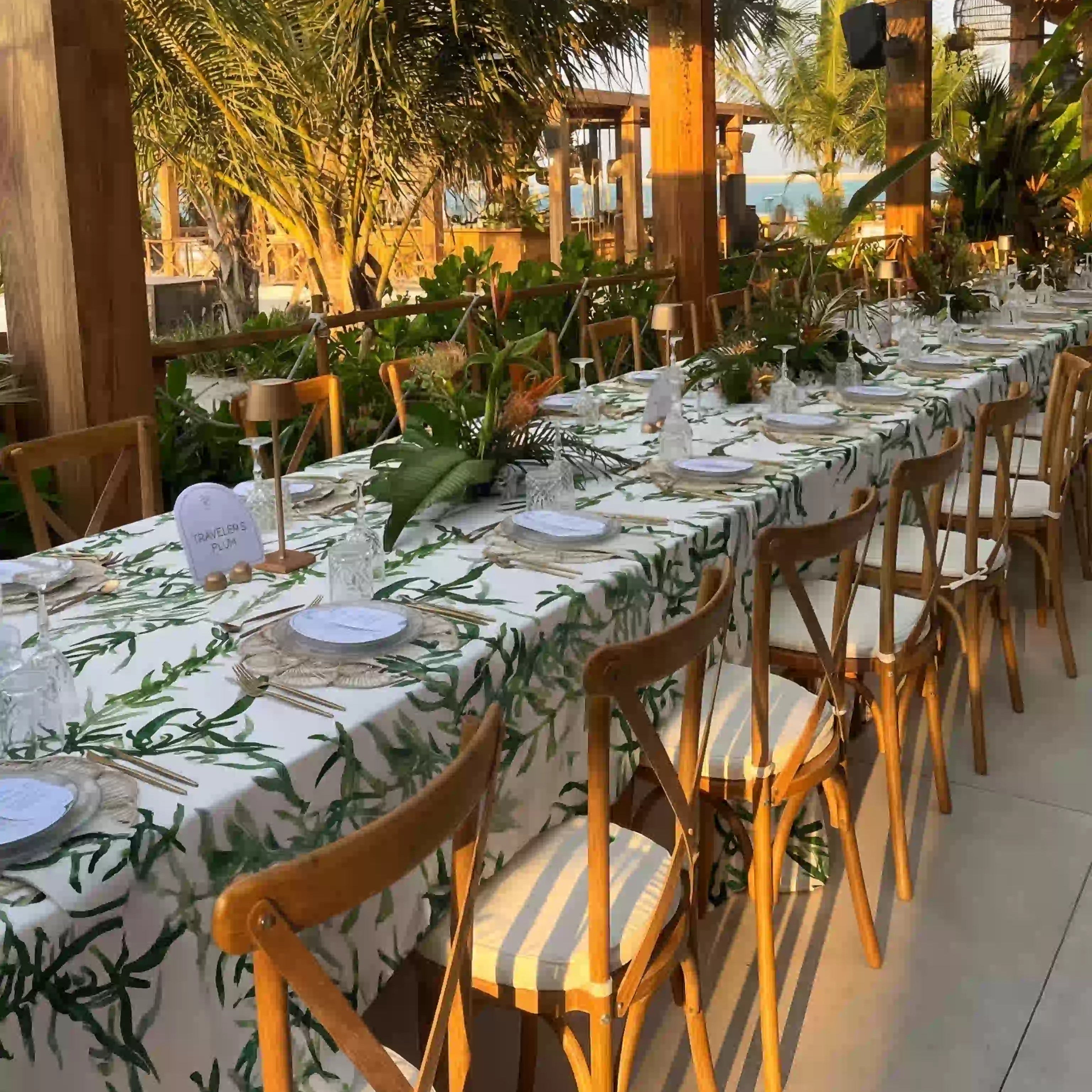 Gardenia Linen Tablecloth: Outdoor dining setup with a white tablecloth featuring green foliage, elegantly set with tableware under a pergola at a beachside event.