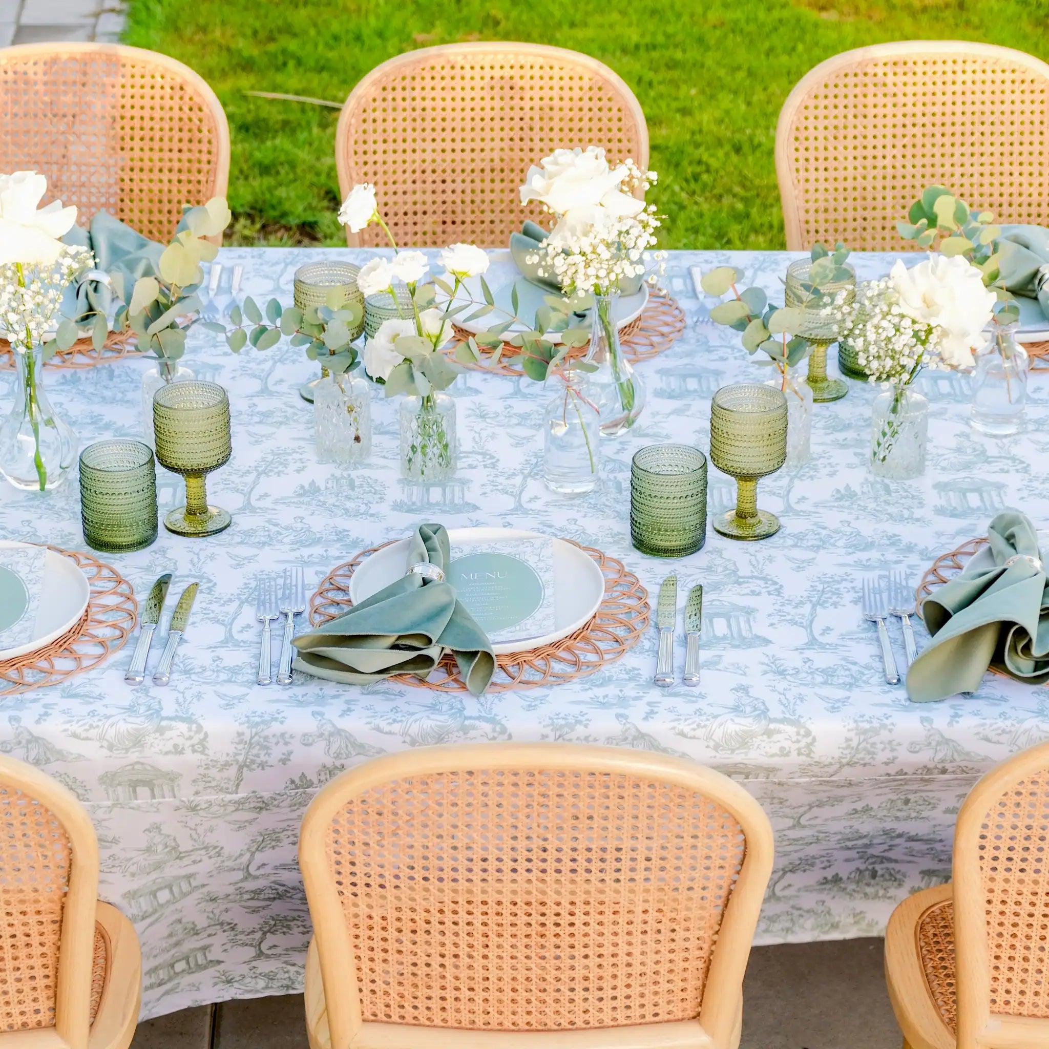 An outdoor dining table set with a white tablecloth featuring a light blue toile-style print, white plates, sage green napkins, green textured goblets, and floral centerpieces, surrounded by woven cane chairs.