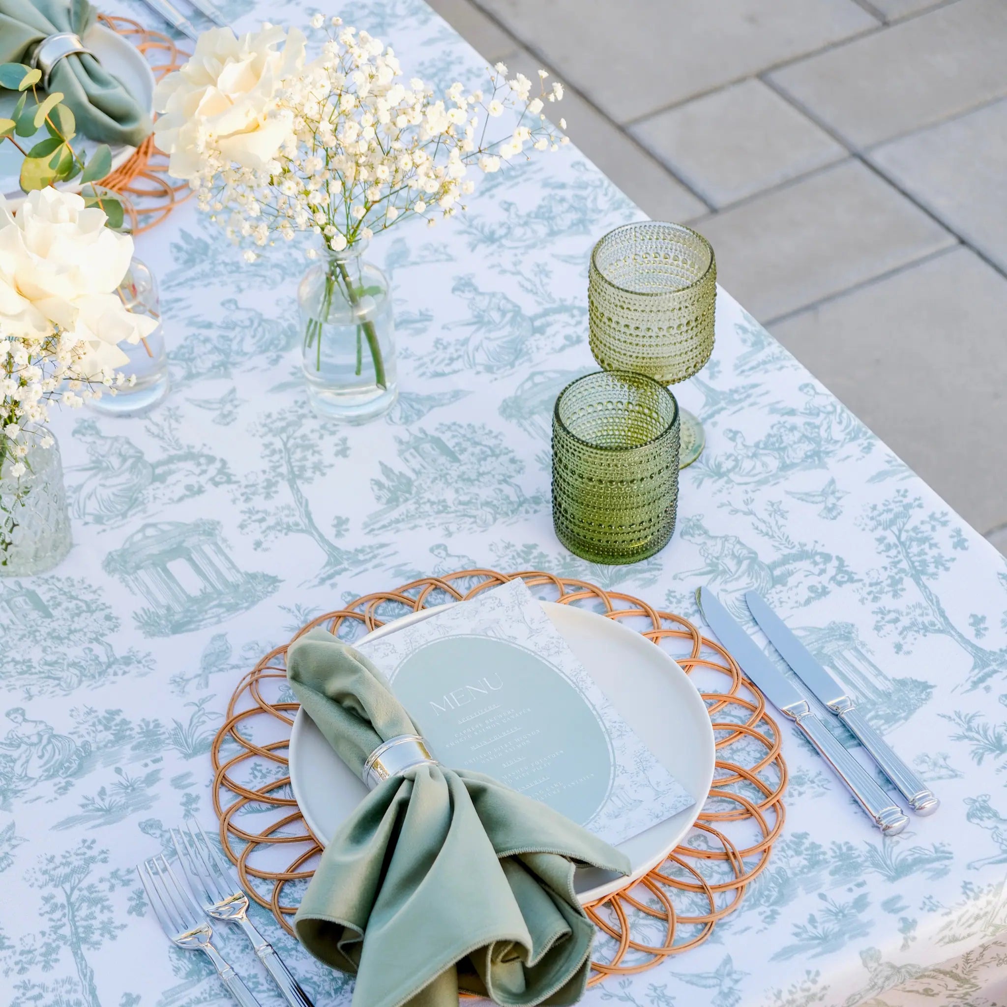 A styled outdoor table setting with a light blue and white toile print tablecloth, white plates, a sage green napkin, silver flatware, and textured green glass tumblers on a stone patio.