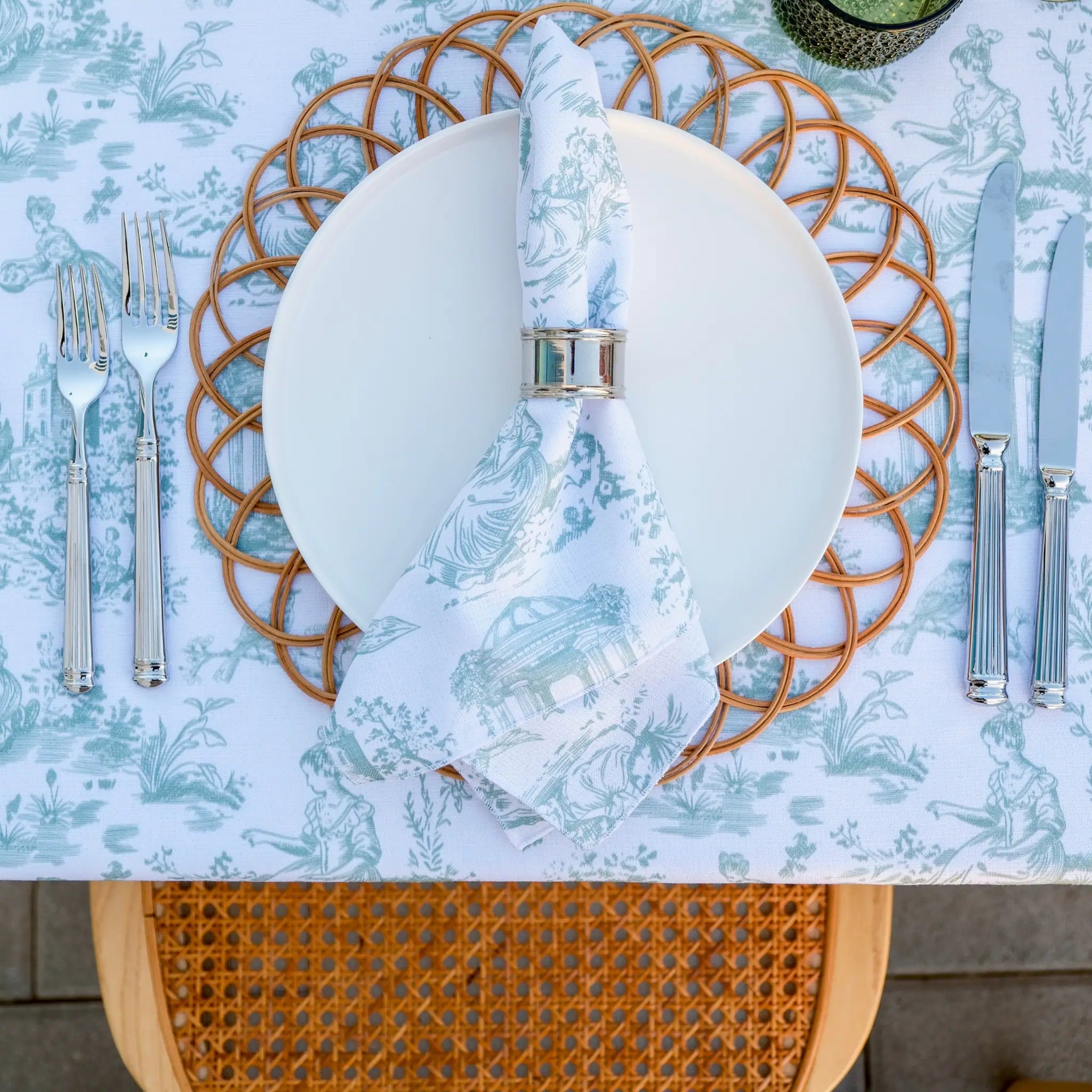 A styled table setting featuring a white ceramic plate on a round woven brown placemat, with a light blue and white toile-print napkin tied in a silver ring. The table is covered in a matching light blue and white toile-patterned tablecloth, with silver forks and knives on either side, and a wooden chair back is visible at the bottom.