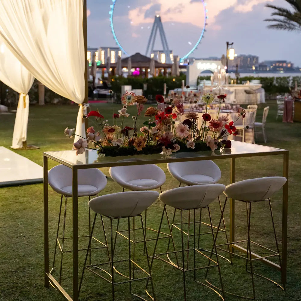 HIGH GLASS RECTANGULAR TABLE W/ GOLD BASE at an elegant outdoor event, surrounded by white bar stools and floral centerpiece, perfect for sophisticated gatherings.