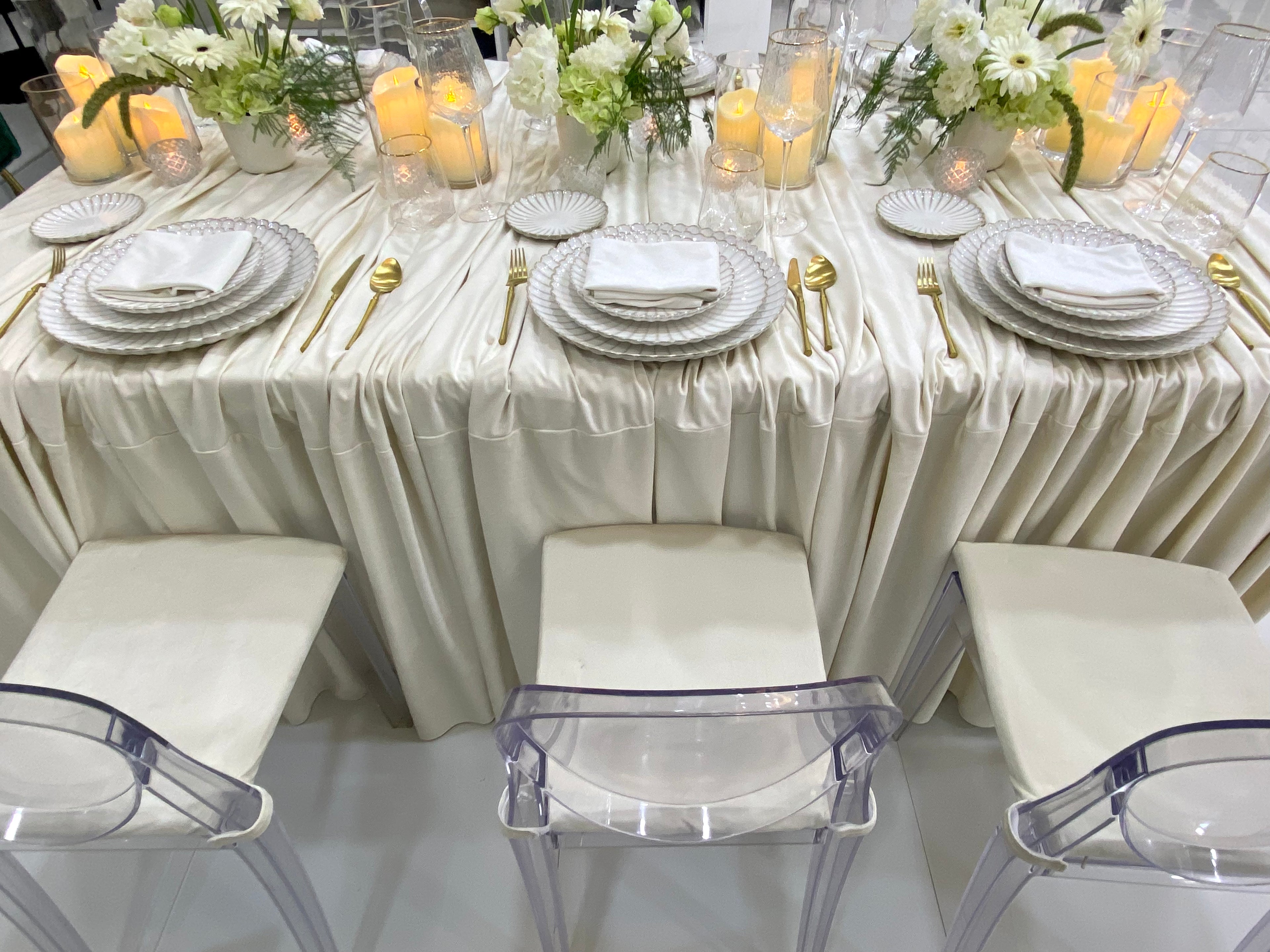 Classic Cream-Dining Setup featuring a banquet table with ivory-satin drape, gold-tone flatware, white floral centerpieces, and acrylic ghost chairs, perfect for elegant events.