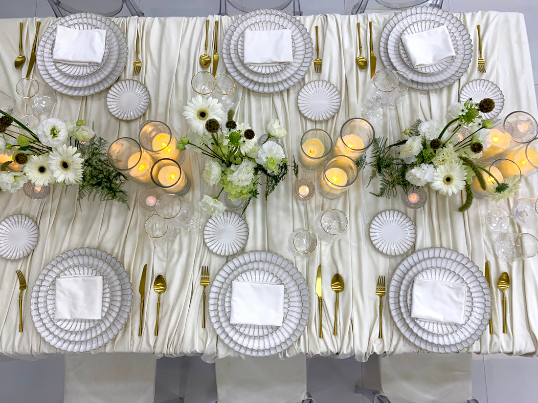 Classic Cream-Dining Setup: Elegantly arranged table for eight, featuring scalloped plates, gold cutlery, glassware, ghost chairs, floral centerpieces, and lit candle vases.