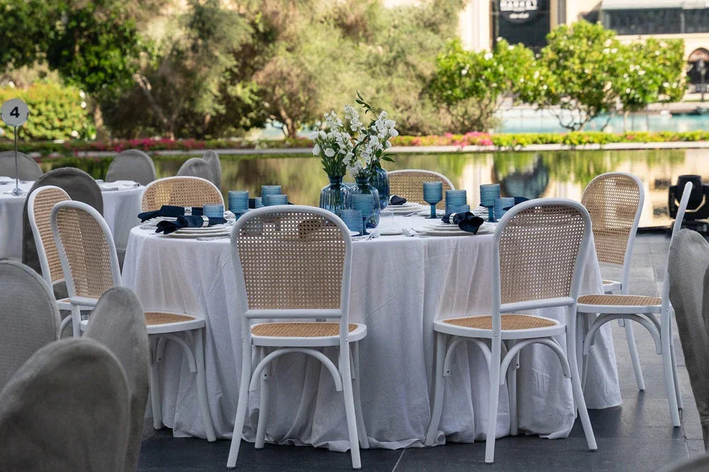 Blue COLORED GLASS VASE on a party table, elegantly holding white flowers amidst an outdoor dining setup with wicker-seated chairs, perfect for enhancing event décor.