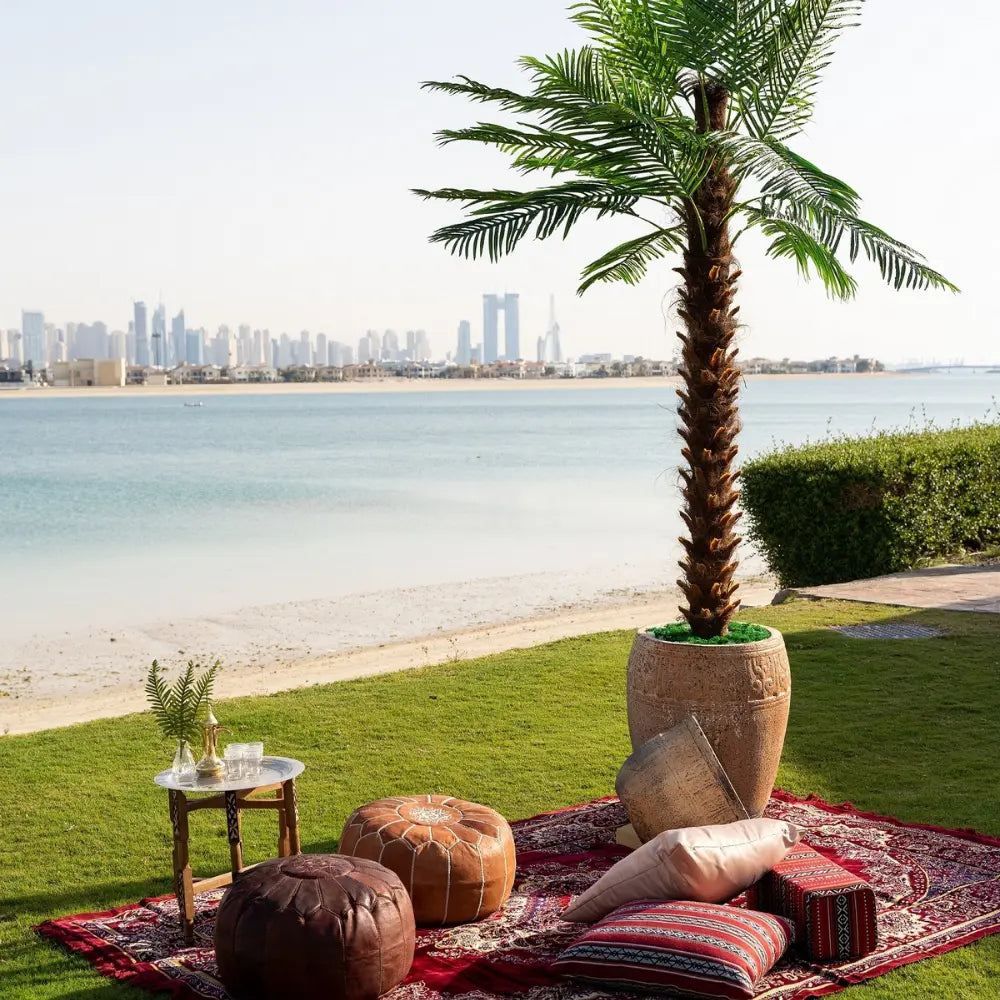 Palm Tree: A 4-meter artificial palm tree enhances an outdoor lounge by the water, creating a tropical, Arabian-style setting with poufs and a rug.
