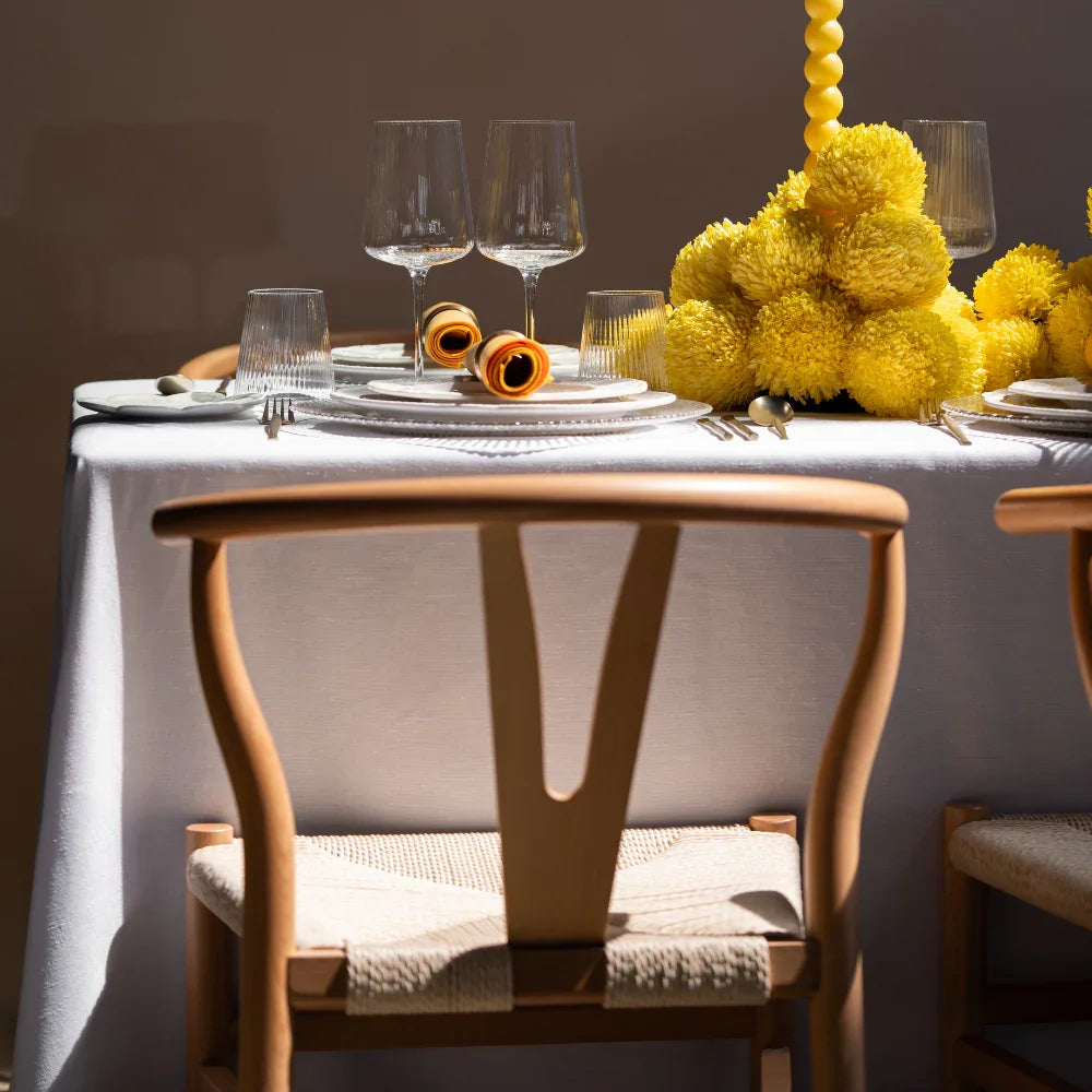 Wishbone Chair with woven seat at elegantly set dining table featuring white plates, yellow-orange napkins, and a yellow floral centerpiece.