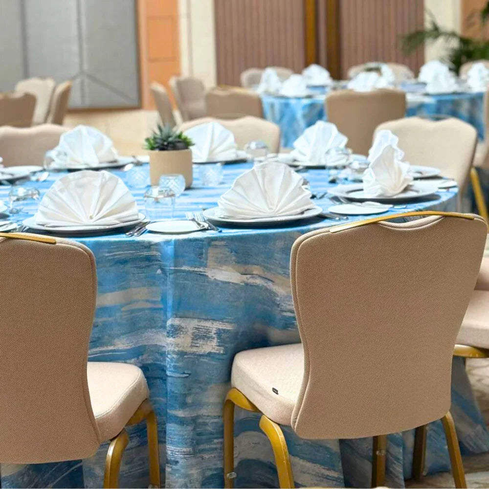 Blue Brushed Round Tablecloth elegantly drapes over a dining table, set for a banquet, enhancing event décor with its sophisticated gold brushed pattern.