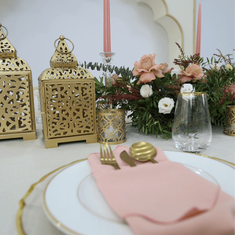 Ramadan Luxe- Dining Setup featuring a table with gold candlesticks, pink napkins, gold lantern, floral centerpiece, and gold cutlery for 8 guests.
