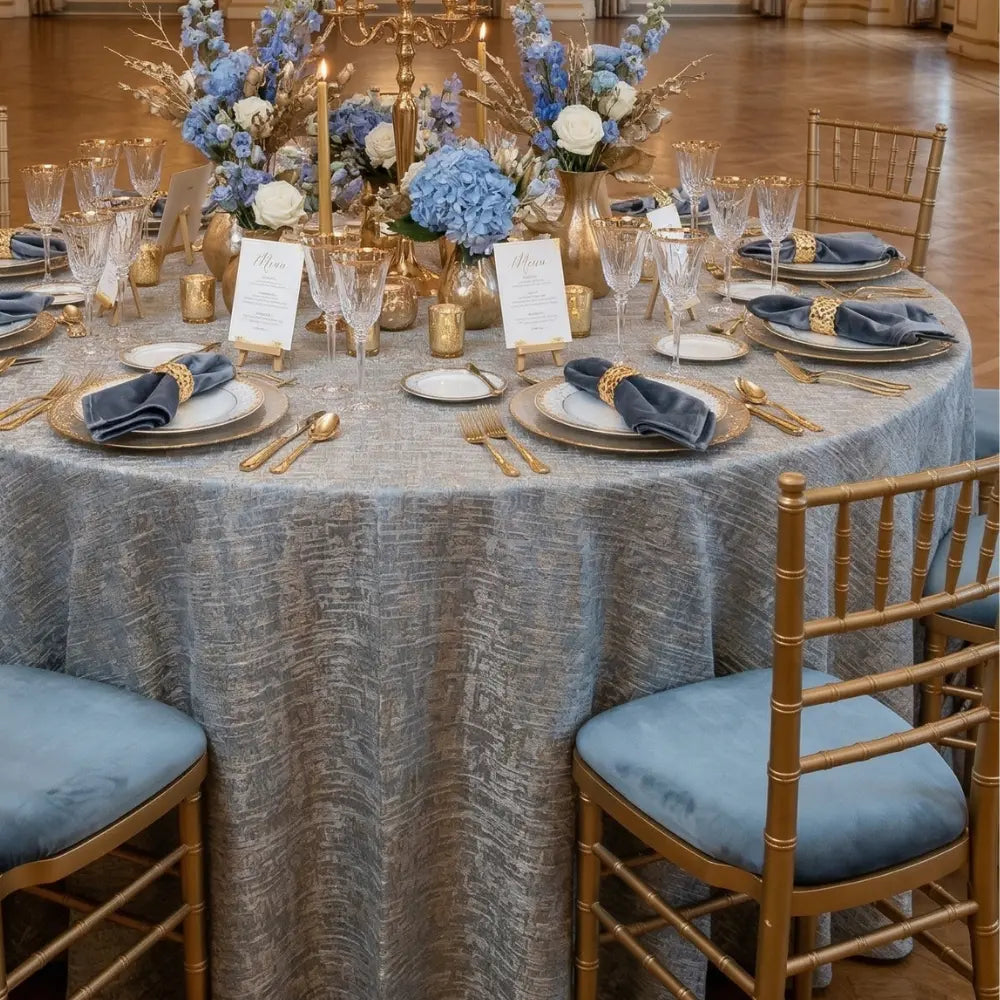 Silver Blue w/ Sand Jacquard Round Tablecloth elegantly draped on a formal table, set with gold-trimmed dinnerware, crystal stemware, and a floral gold centerpiece.