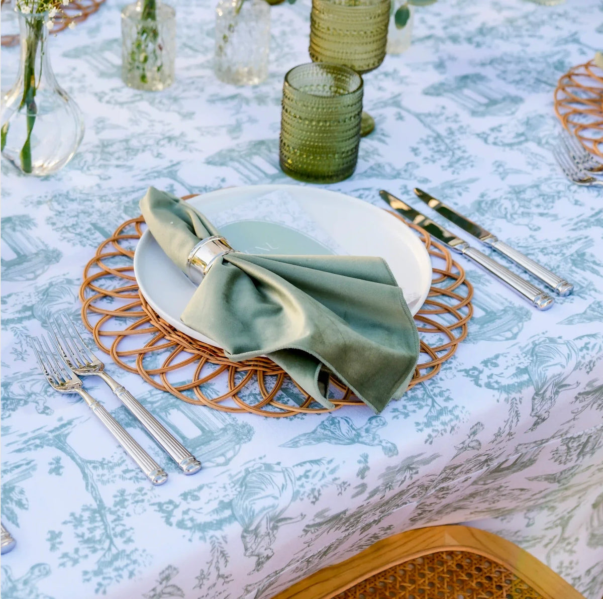 A styled table setting featuring a white tablecloth with a light blue toile de Jouy pattern, a white charger plate, a woven round gold charger, and a sage green satin napkin tied with a gold ring.