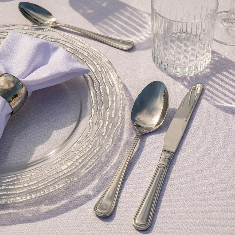 CUTLERY SIMPLE SILVER set elegantly displayed with a charger, napkin, and glass tumbler, showcasing polished stainless-steel craftsmanship ideal for formal dining settings.