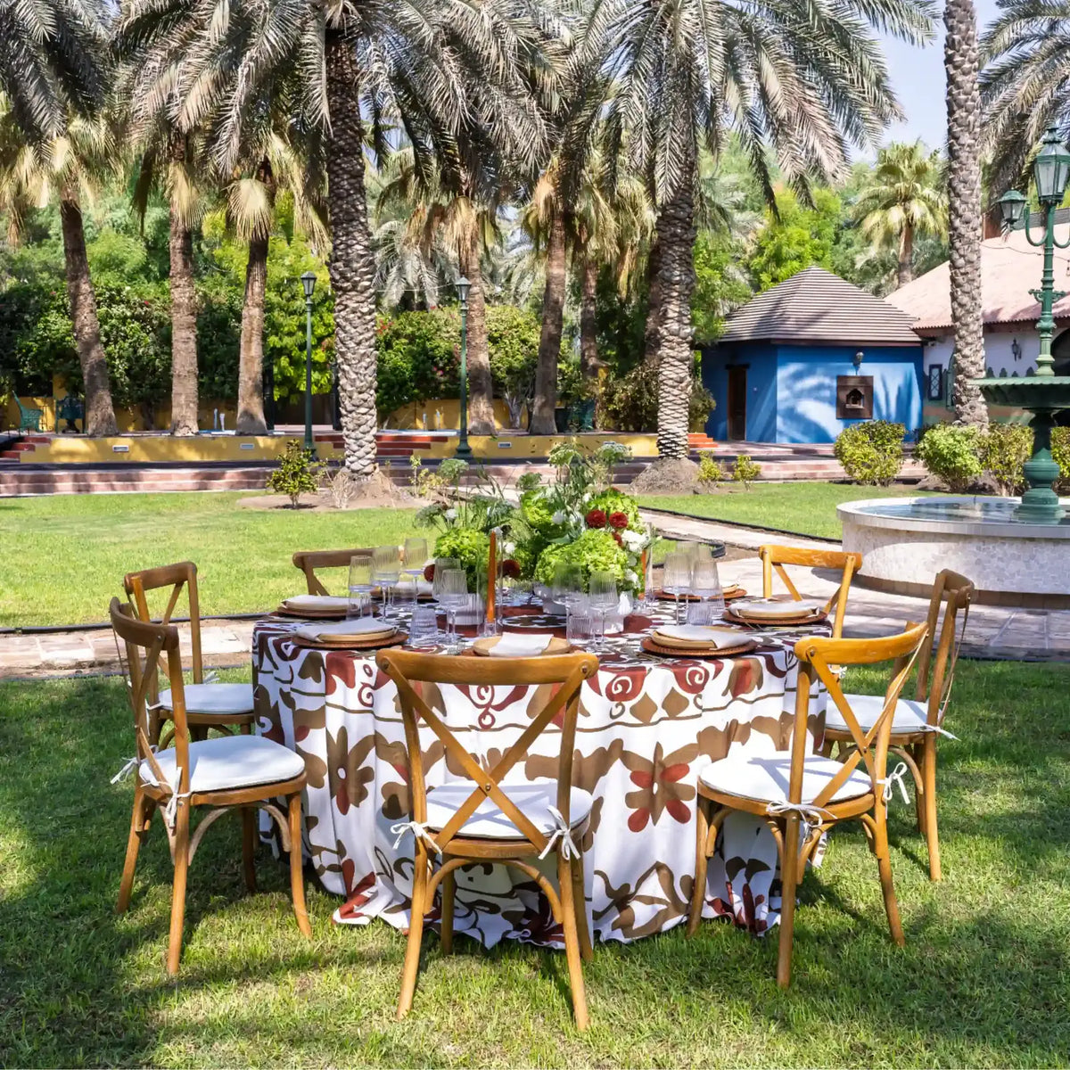 An outdoor dining setup in a lush garden with palm trees, featuring a round table draped in a brown and cream patterned tablecloth, surrounded by six wooden chairs with white cushions. The table is set with glassware, plates, and a floral centerpiece.