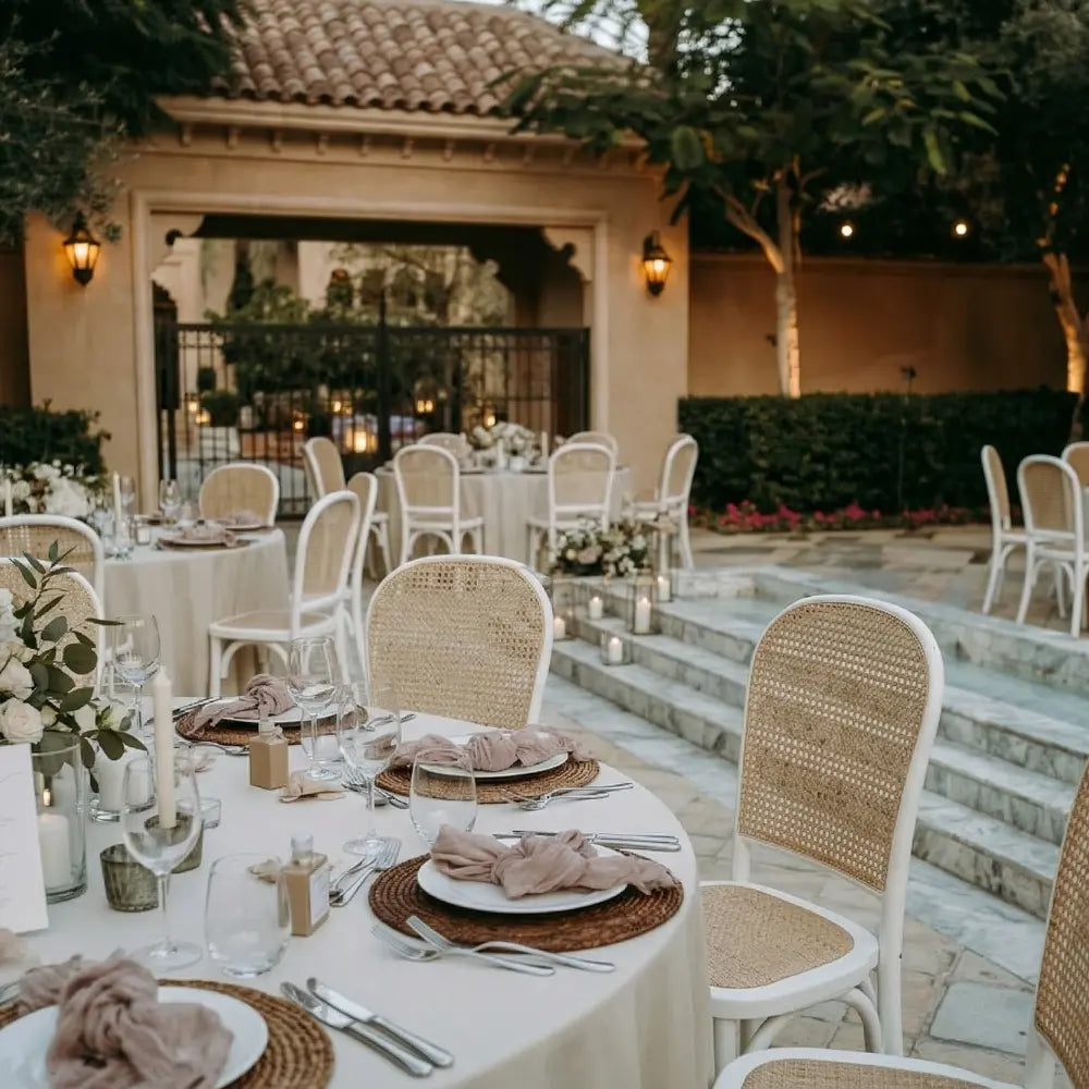 RATTAN CHAIRS arranged around elegantly set tables with ivory linens, enhancing the sophisticated, old-world charm of an outdoor event space.