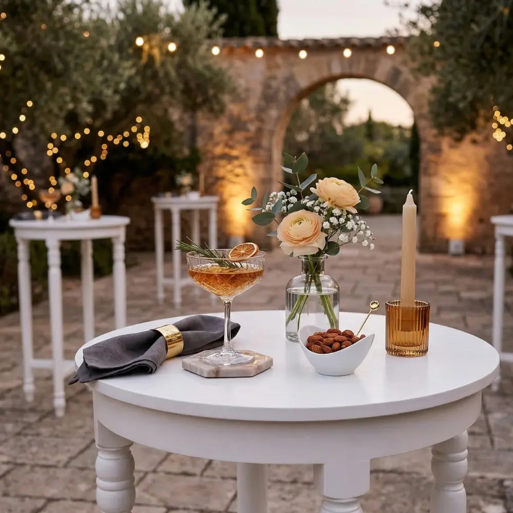 White wood cocktail table set with a cocktail, linen napkin, nuts, floral arrangement, and candle, creating an elegant outdoor event atmosphere.