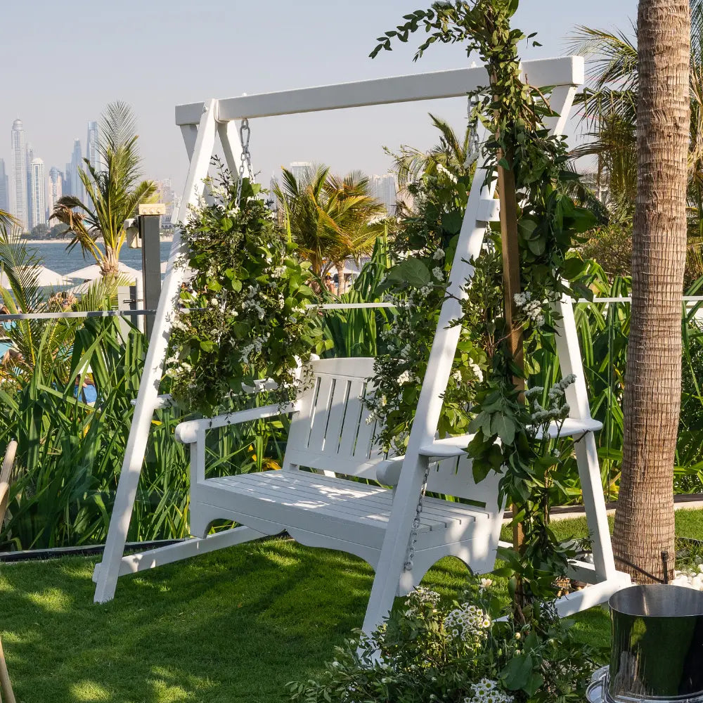 White wooden swing with greenery in a garden setting with palm trees and city skyline.