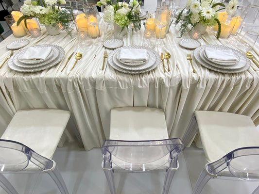 Classic Cream-Dining Setup featuring a banquet table with ivory-satin drape, gold-tone flatware, white floral centerpieces, and acrylic ghost chairs, perfect for elegant events.