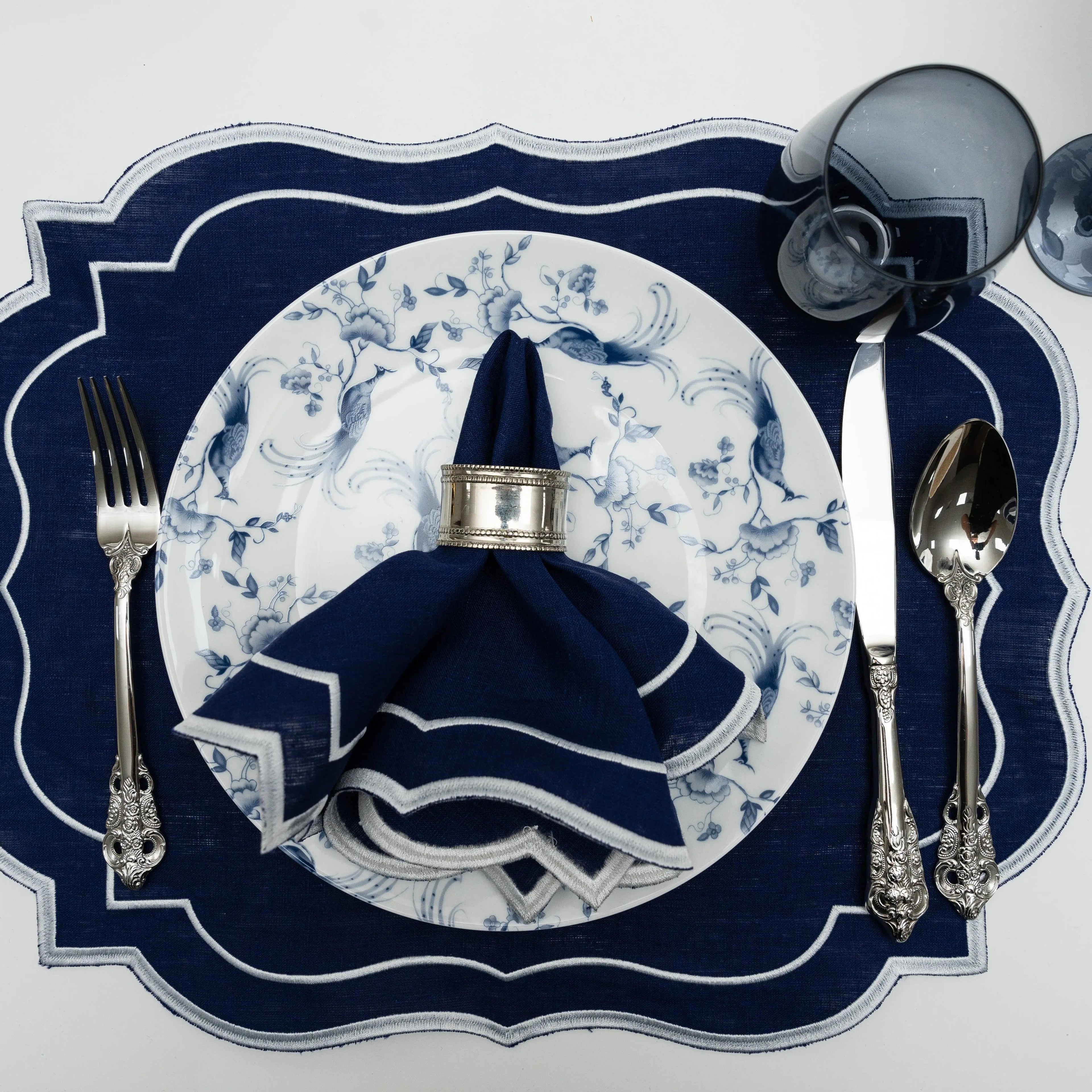 Top-down view of Navy Scalloped Linen Placemats in an elegant table setting with matching napkin, silver cutlery, and a smoky glass goblet.