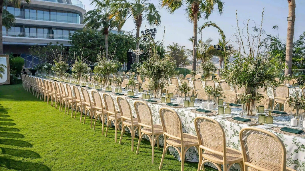 Outdoor event setup with long table and chairs on a grassy area with palm trees in the background.