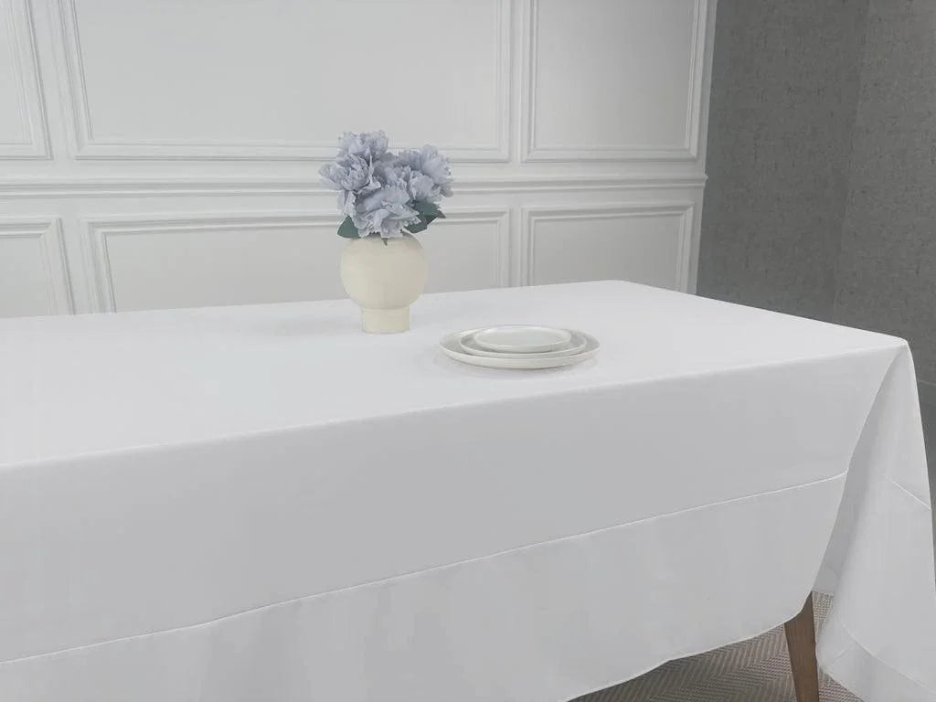 A white table with a vase of flowers, showcasing the Polycotton Tablecloth from Party Social. Perfect for any event, this lightweight tablecloth adds a cloth feel without the need for paper or plastic. Easy to wash and available in various colors and sizes, it&
