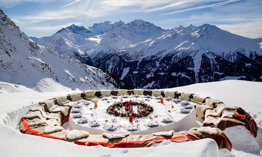 A round, snow-carved dining table set outdoors atop a snowy mountain, surrounded by cushioned seating, with place settings and a festive centerpiece against a panoramic alpine backdrop.