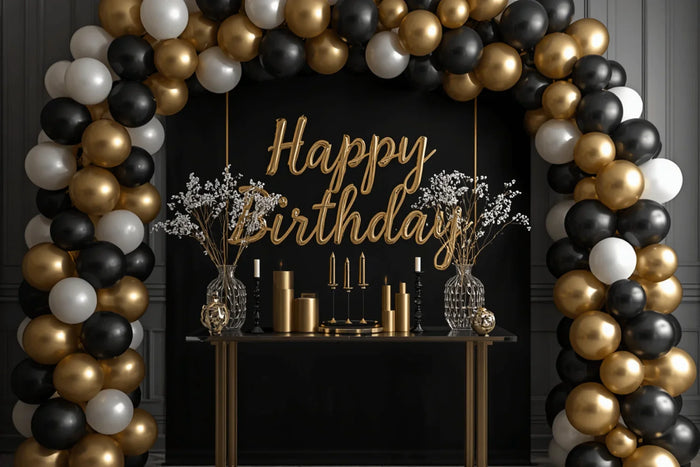 birthday party setup at home with a full balloon arch in matte black, metallic gold, and white, elegant black backdrop with gold lettering