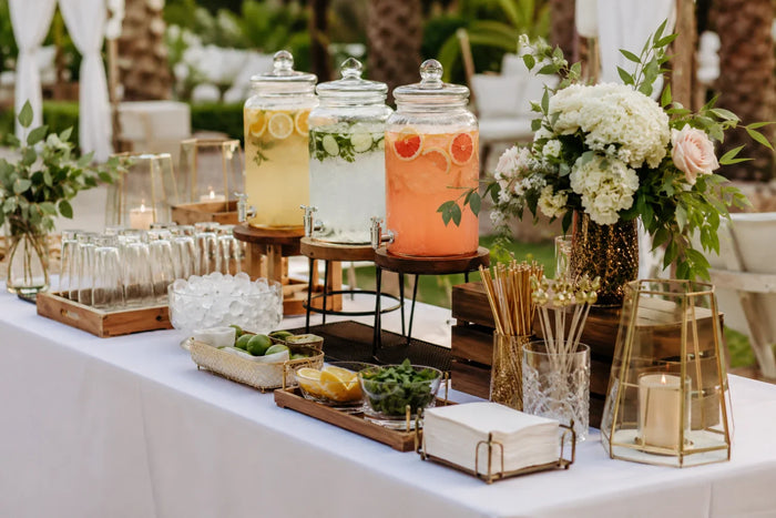 Stylish self-serve drink station with beverage dispensers, glassware, floral arrangements, garnishes, and decorative accents for an elegant outdoor event.