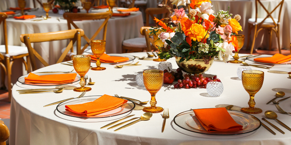 A dinner party table set with plates, glasses, orange napkins on white plates, and a floral centerpiece.