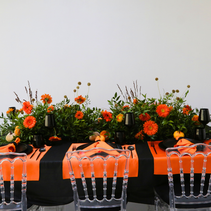 Pumpkins &amp; Petals- Dining Setup with a black tablecloth, orange accents, clear chairs, and a vibrant floral centerpiece, perfect for an elegant dinner party for eight.