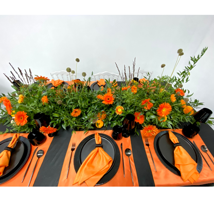 Pumpkins &amp; Petals- Dining Setup: Elegant tablescape featuring lush foliage, vibrant orange flowers, black dinnerware, and modern flatware, set for eight guests with sleek white chairs.