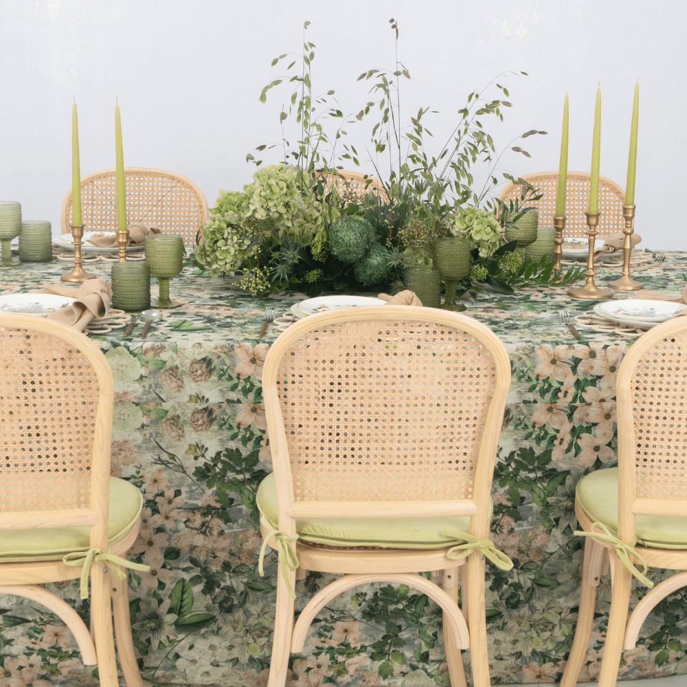 Garden Bliss- Dining Setup featuring a table set for eight with rattan chairs, plates, gold cutlery, goblets, candle holders, and a flower centerpiece.