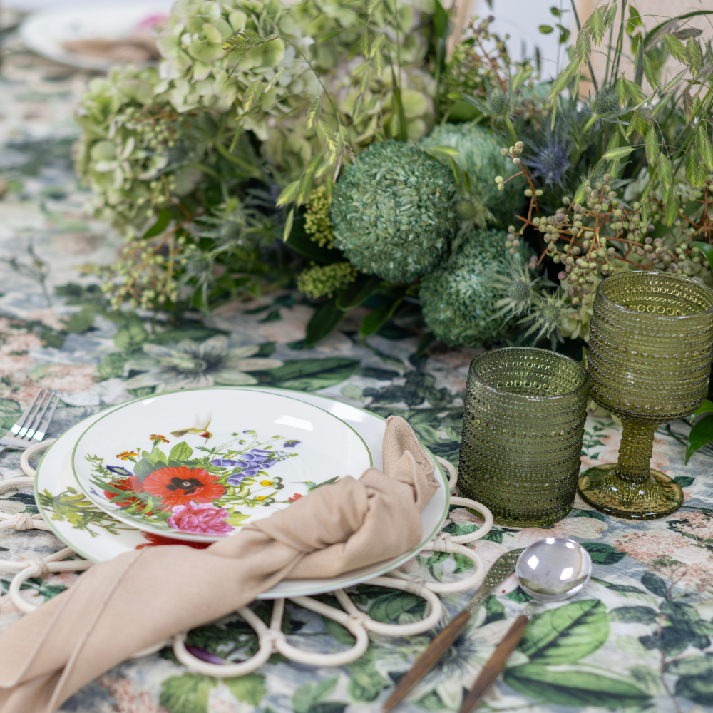 Garden Bliss- Dining Setup featuring a table with plates, glasses, gold cutlery, napkins, rattan chairs, flower centerpiece, and candle holders for 8 guests.