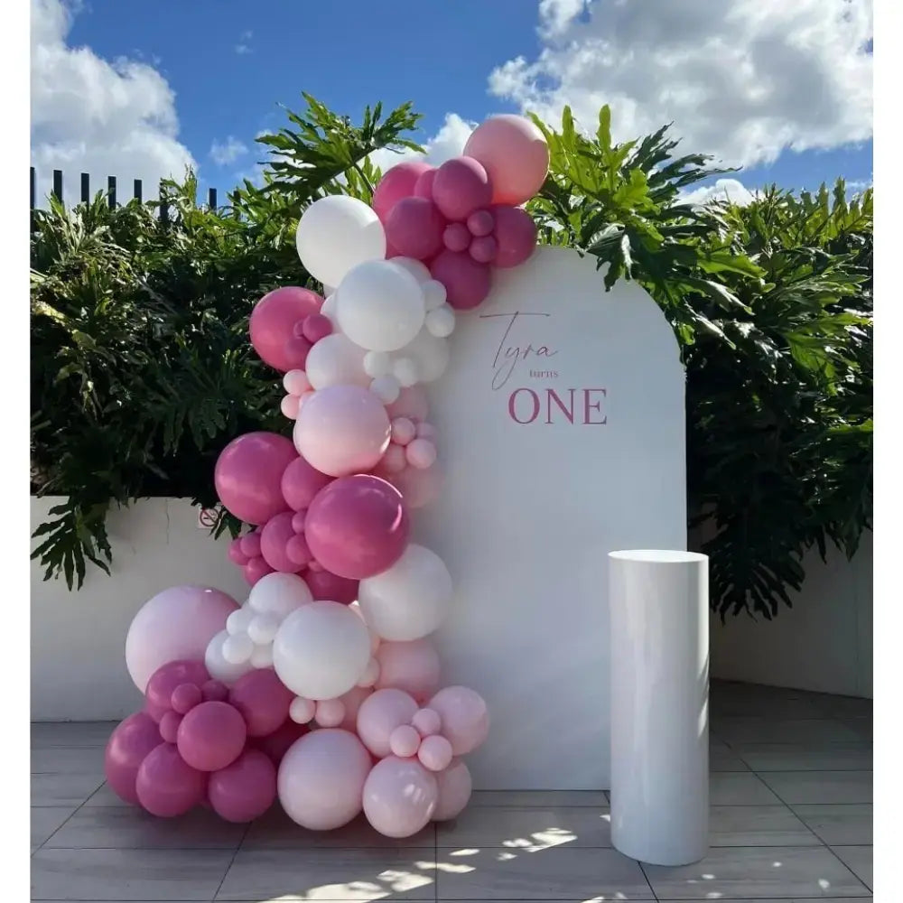 One-derful Blush Setup featuring a white arched backdrop with “Tyra turns ONE,” pink and white balloon garland, and a white pedestal.
