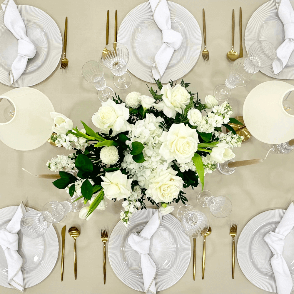 Sand Elegance- Dining Setup features a table set for eight with white plates, gold cutlery, glasses, napkins, ivory chairs, lamps, and a floral centerpiece.