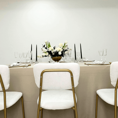 Sand Elegance- Dining Setup featuring a table with a flower centerpiece, surrounded by simple ivory chairs, gold cutlery, and elegant tableware for eight guests.