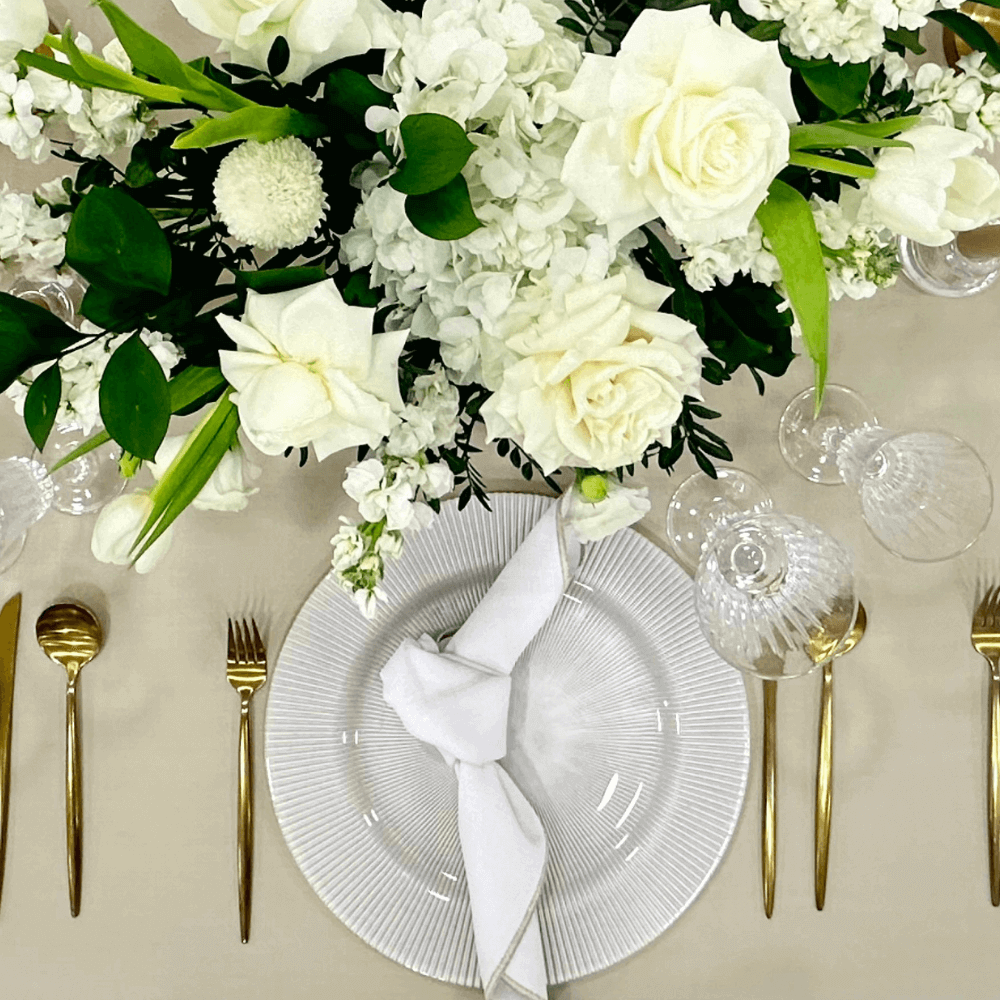 Sand Elegance- Dining Setup featuring a table with a flower centerpiece, gold cutlery, plates, glasses, and napkins arranged for eight guests.